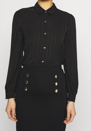 Žena oblečená v černé košilové blůze s jemnými pruhy a v černé sukni s vysokým pasem, která má dva řady zlatých knoflíků.