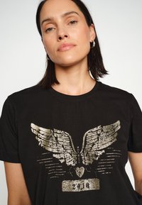 Melns kokvilnas T-krekls ar metālisku zelta spārnu dizainu un gadu "2010" zem tā, uz gludas tekstūras.