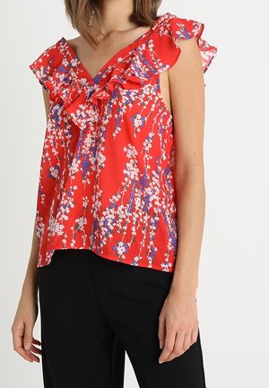 Femme portant un haut sans manches rouge à fleurs avec un col à volants, associé à un pantalon noir, sur fond blanc.