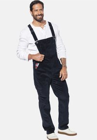 Marineblaue Corduroy-Latzhose mit verstellbaren Trägern, featuring eine Fronttasche mit einem Karo-Akzent, ergänzt durch ein weißes Langarmshirt und Sneaker.