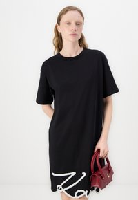 Černé bavlněné šaty ve stylu trička s krátkými rukávy, na spodní části s bílým kurzivním textem. Modelka drží malou bordó kabelku.