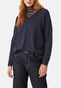 Felpa blu navy con scollatura a V e spalle scese, realizzata in tessuto morbido, indossata sopra un dolcevita a righe multicolori, abbinata a jeans scuri.