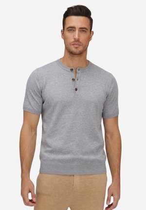 Mann mit dunklem Haar, der ein hellgraues kurzärmeliges Henley-Shirt und beige Hosen trägt, steht vor einem schlichten Hintergrund.