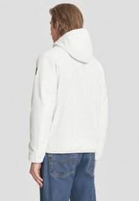 Giacca bianca con cappuccio e texture liscia, dotata di un logo visibile sulla manica sinistra e una vestibilità rilassata. Indossata con jeans in denim blu.