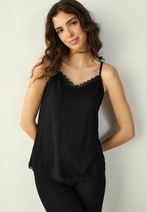 Ung kvinde med langt, bølget brunt hår iført en sort ærmeløs top med blonder, stående mod en ensartet lys baggrund.