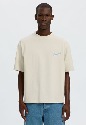 Mand iført en ensfarvet, offwhite kortærmet T-shirt med lyseblå tekst på venstre bryst og blå jeans, stående mod en neutral baggrund.
