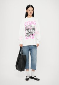 Sudadera blanca de manga larga con un diseño gráfico en negro y rosa, combinada con unos jeans recortados de color azul claro y un bolso tote negro.