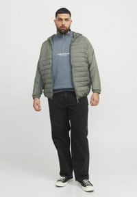 Olivgrön puffjacka med dragkedja fram, lagd över en blå sweatshirt. Svarta byxor och sneakers kompletterar outfiten.