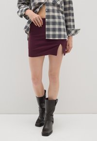 Mini gonna bordeaux con spacco laterale, abbinata a una camicia oversize a scacchi bianca e nera. Modella che indossa stivali neri al ginocchio.