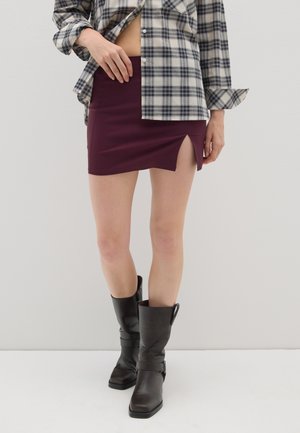 Minifalda burdeos con una abertura lateral, combinada con una camisa de cuadros oversized en blanco y negro. Modelo con botas negras hasta la rodilla.
