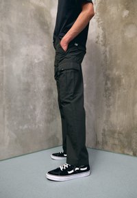 Man som bär svarta cargo-byxor och svarta-och-vita sneakers, stående mot en texturerad grå betongvägg och golv.