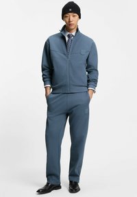 Man wearing blue zip-up jacket and matching pants with hands in pockets, striped shirt, tie, black beanie, and black shoes.