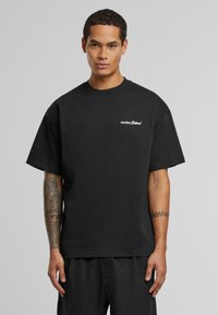 Sort bomulds T-shirt med afslappet pasform, der har korte ærmer og et hvidt logo på venstre bryst. Glat tekstur, enkelt design.