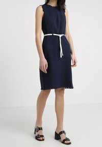 Námorně modré šaty bez rukávů s texturovanou tkaninou, se třepivým lemem a bílým opaskem na zavázání. Černé sandály na podpatku s přezkami.
