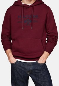 Bordeaux hoodie med känguruficka, dragsko på huvan och tryckt logo i marinblått. Tillverkad i bomull med ribbad manschett och nederkant. Enkelt design.