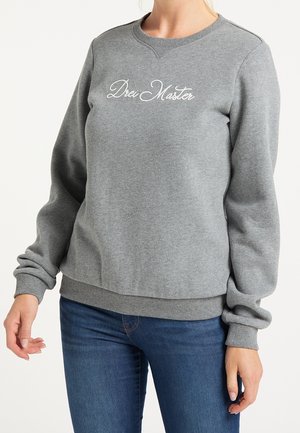 Kvinde iført en grå crewneck-sweatshirt med teksten "Drei Master" og blå jeans, stående med afslappede arme mod en hvid baggrund.