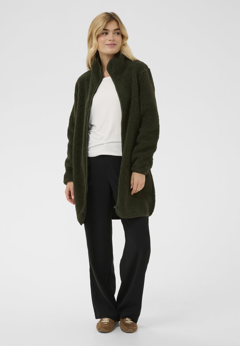 Grüner, flauschiger Fleece-Mantel mit hohem Kragen, offenem Vorderteil und langen Ärmeln, kombiniert mit schwarzen Hosen und braunen Freizeitschuhen.