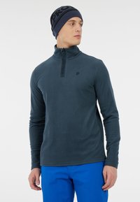 Marineblå fleece pullove med halvlukning og høj krave. Har et diskret logo og en glat tekstur. Bæres med klare blå bukser.