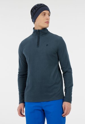 Granatowy polarowy sweter z pół-zamkiem i wysokim kołnierzem. Posiada subtelne logo i gładką fakturę. Noszony z jasnoniebieskimi spodniami.