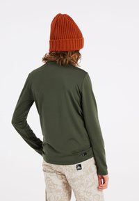 Camisa de manga larga verde oliva con una textura suave, combinada con pantalones beige con estampado. Un gorro de punto naranja completa el conjunto.