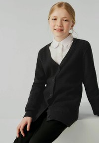Cardigan nero lavorato a maglia con scollatura a V, caratterizzato da un motivo testurizzato, maniche lunghe e cinque bottoni. Indossato su una camicia bianca con colletto.