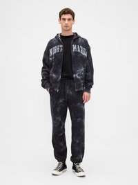 Νεαρός άνδρας φορώντας μαύρη φούτερ με σχέδιο tie-dye και αντίστοιχες φόρμες με την ένδειξη "NIGHTMARE", μαύρο πουκάμισο και μαύρα αθλητικά παπούτσια ψηλής κορυφής, όρθιος.