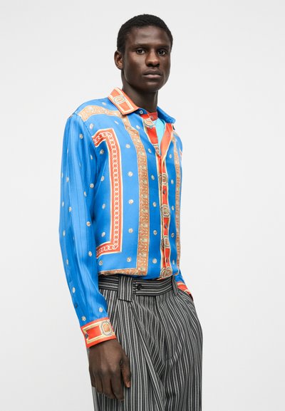 Bărbat purtând o cămașă vibrantă cu mâneci lungi, albastră și portocalie, cu detalii aurii și pantaloni cu dungi verticale alb-negru.