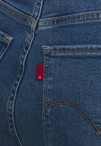 Siniset farkut, joissa on keltainen kontrastisaumaus, takataskussa punainen merkki ja tunnistettavat käyrät taskusaumadetaljit.