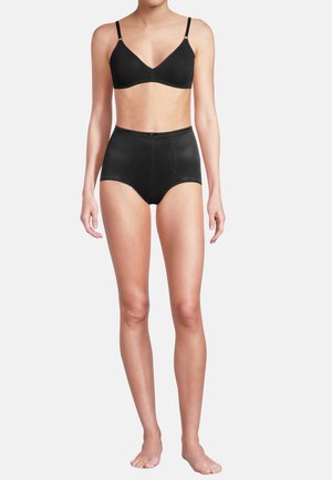 Vrouw staat op blote voeten, draagt een zwarte bh en zwarte, hoog uitgesneden ondergoed tegen een eenvoudige witte achtergrond.