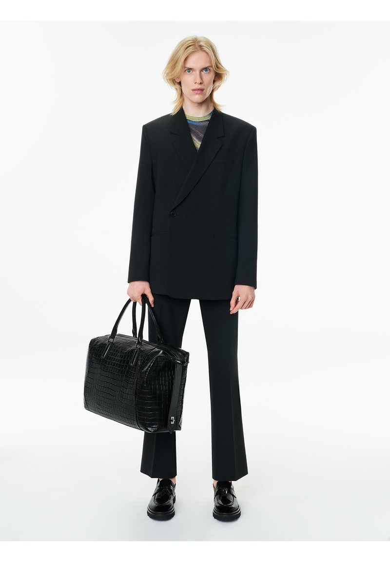 Blazer negro de doble botonadura sobre una camisa a rayas, combinado con pantalones negros ajustados y zapatos negros brillantes. Sosteniendo un bolso negro texturizado.