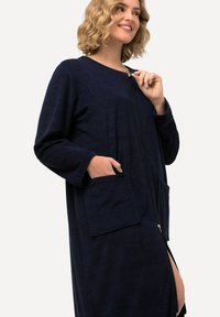 Námořnický modrý župan na zip vyrobený z měkké tkaniny, s dlouhými rukávy, dvěma předními kapsami a bočním rozparkem pro snadný pohyb.