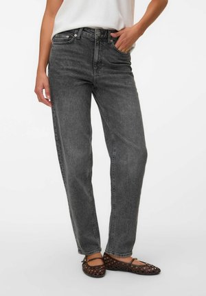 Vrouw draagt zwarte, gewassen, rechte jeans met hoge taille, wit topje, één hand in de zak en donkerbruine gevlochten platte schoenen.
