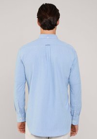 Camisa abotonada de color azul claro con rayas verticales blancas, mangas largas, cuello tipo polo y detalle de pliegue en la parte posterior.