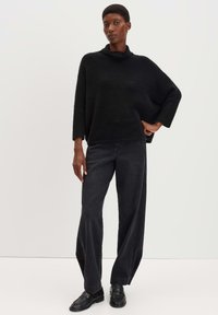 Schwarzer, strukturierter oversized Pullover mit trichterförmigem Ausschnitt, kombiniert mit locker sitzenden schwarzen Jeans und schwarzen Slippern.