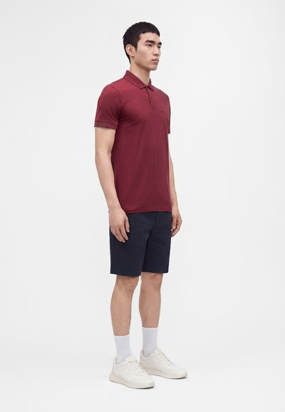 Jeune homme portant un polo bordeaux, un short bleu marine, des chaussettes blanches et des baskets blanches, debout devant un fond blanc uni.