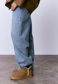 Botines de ante color canela con costuras decorativas, combinados con jeans de mezclilla azul claro. Los jeans tienen un corte relajado y costuras visibles.