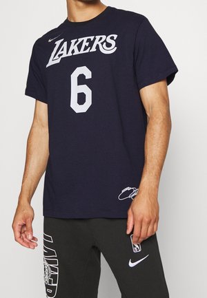 Mężczyzna ubrany w czarną koszulkę Lakers z numerem 6 i czarne spodnie z logo NBA i Nike, stojący na jednolitym tle.
