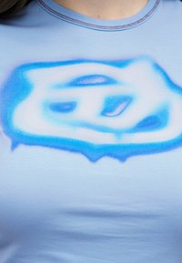 Ανοιχτό μπλε πουκάμισο με μπλε και λευκό αφηρημένο σχέδιο ψεκασμού χρώματος κατά μήκος του στήθους και στρογγυλή λαιμόκοψη κοντά στον λαιμό.