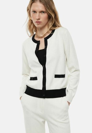 Donna che indossa un cardigan bianco con rifiniture nere e bottoni, pantaloni bianchi, una maglietta nera e una collana a catena d'oro, posando con le mani nelle tasche.