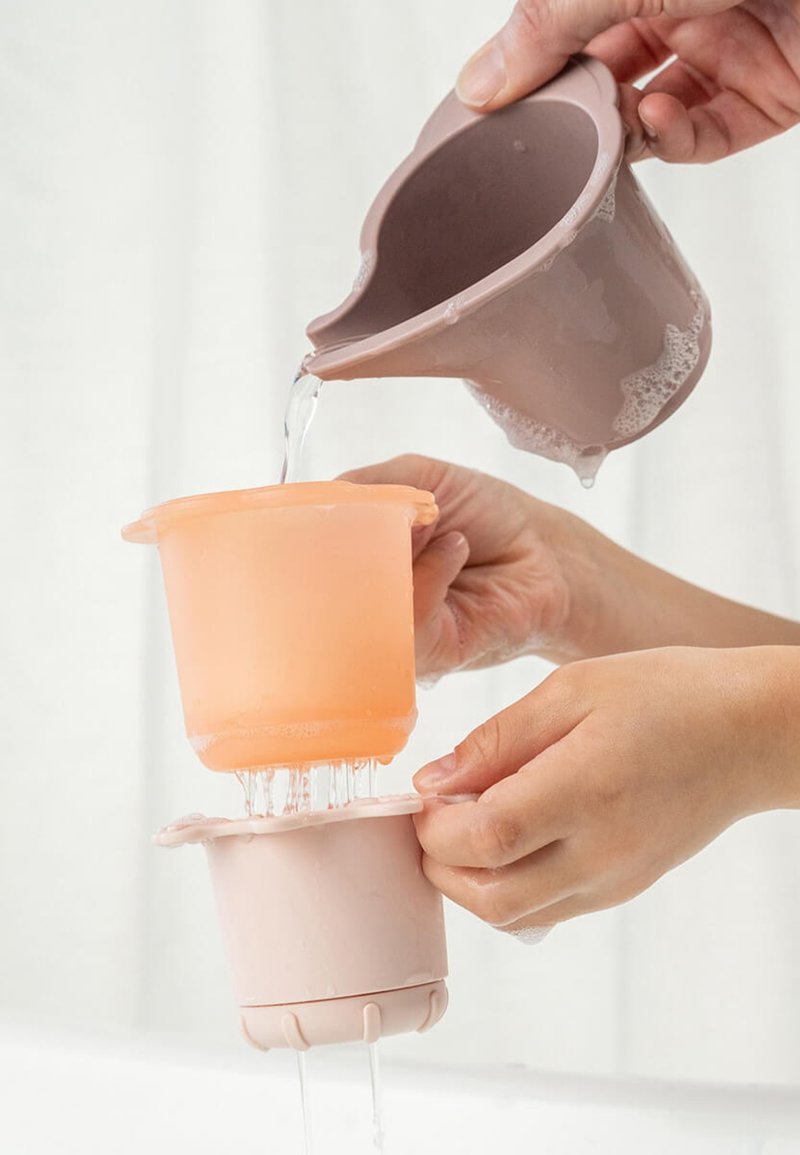 Handen gieten water uit een mauve container in een oranje zeef boven een lichtroze beker, terwijl het water door de gaatjes wegstroomt.