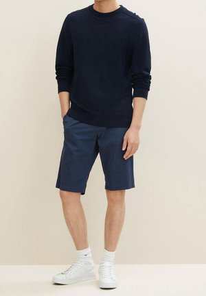 Mann trägt marineblaues Sweatshirt mit langen Ärmeln, blaue gemusterte Shorts, weiße Socken und weiße Turnschuhe, steht mit einer Hand in der Tasche.