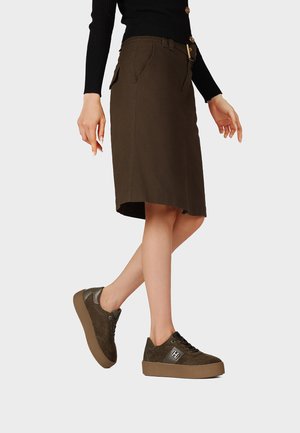 Donna che indossa una gonna marrone scuro al ginocchio con tasca laterale e sneakers marroni con plateau abbinate, su uno sfondo chiaro e semplice.