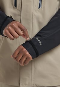 Giacca impermeabile beige con maniche nere dotata di regolazione a strappo ai polsini. Tessuto anti-strappo, logo visibile sulla manica.