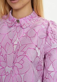 Bluză florală violetă cu guler căptușit, modele complicate de petale, țesătură texturată și un nasture accent în formă de inimă la guler.