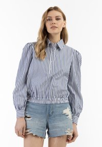 Stribet skjorte i blå og hvid med lange, puffede ærmer og elastisk talje, parret med slidte denimshorts.