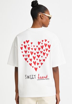 Femme portant un T-shirt blanc avec un groupe de cœurs rouges formant une grande forme de cœur et le texte "SWEET heart" dans le dos.