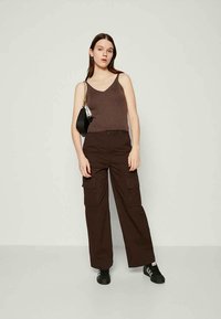 Donna in piedi che indossa un top senza maniche marrone, pantaloni cargo marrone scuro, sneakers nere e una piccola borsa a tracolla nera con una catena.
