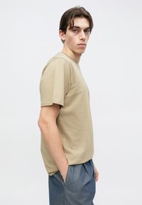 Μπεζ βαμβακερό T-shirt με κοντά μανίκια, στρογγυλή λαιμόκοψη και άνετη εφαρμογή. Φορεμένο με μπλε παντελόνια, παρουσιάζοντας έναν μινιμαλιστικό σχεδιασμό.