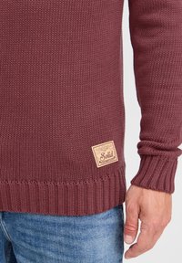 Burgunderfarbener gestrickter Pullover mit gerippten Bündchen und Saum, der ein strukturiertes Muster und ein Lederlogo-Tag an der unteren Seite aufweist.