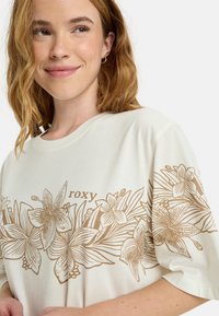 Off-white t-shirt featuring a gold floral design and the word "roxy." The fabric is smooth with short sleeves and a crew neckline.